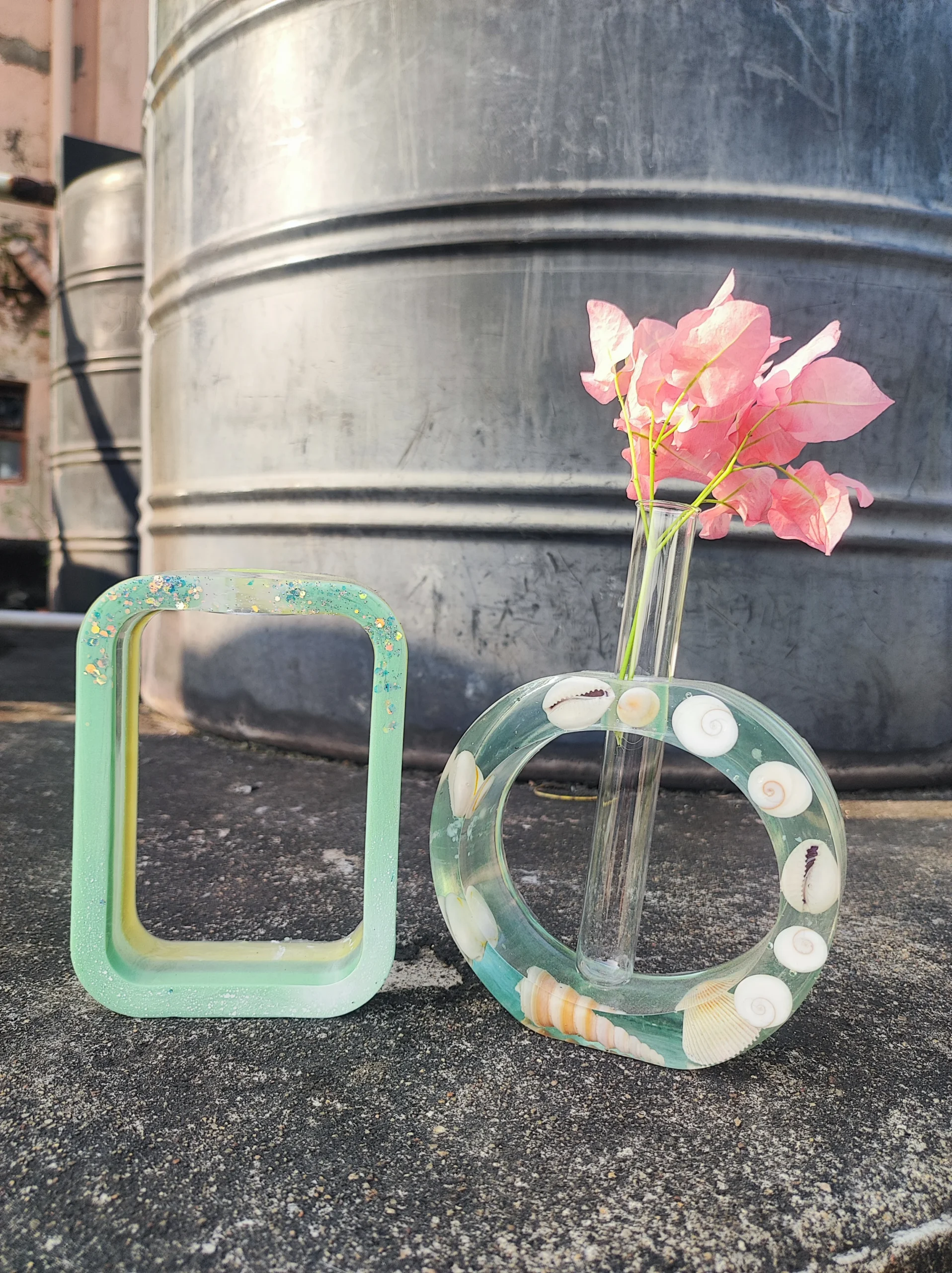 Pink bougainvillea flowers in a tall glass vase placed on the asphalt beside a green, shell-decorated circular sculpture, with a large metal tank in the background. - Bling On Decor® | Luxury Handmade Home Decor ANJU BALA
