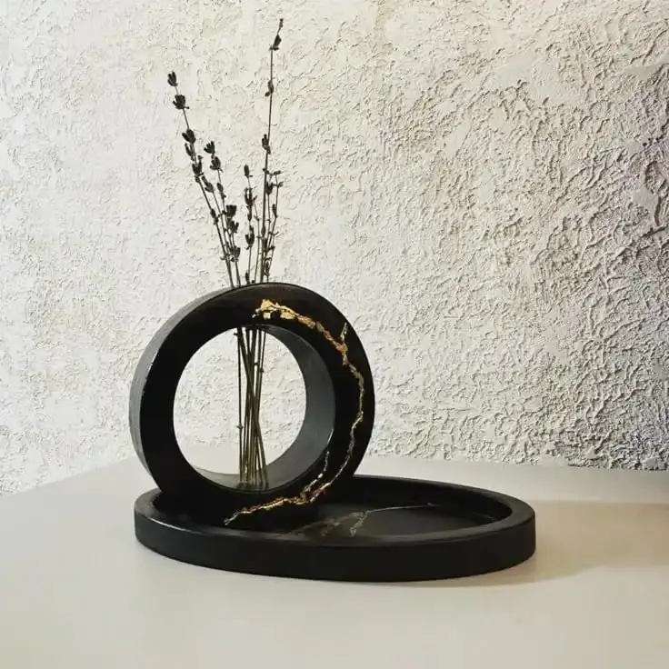 🖤-Handmade-Luxury-Black-Resin-Kintsugi-Ring-Vase-with-Tray.webp - Bling On Decor® | Luxury Handmade Home Decor Black circular sculpture with a hollow ring and a vase of dried stems, resting on a round base against a textured wall. - Bling On Decor® | Luxury Handmade Home Decor ANJU BALA