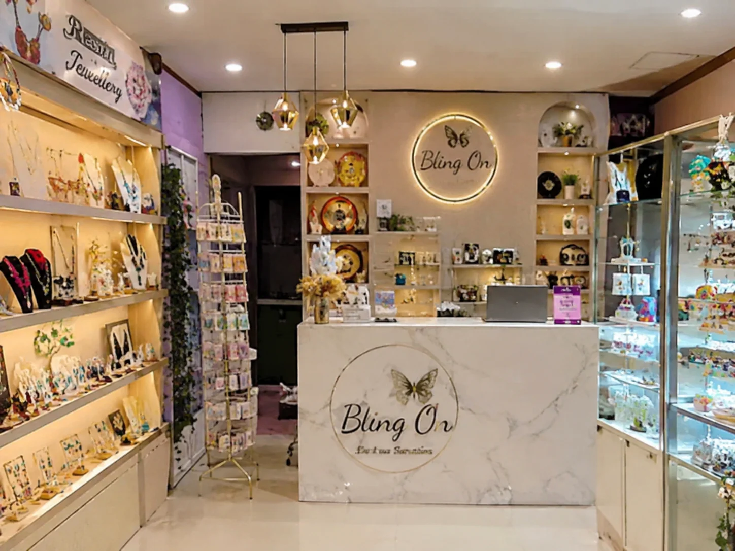 Inside a bright jewelry shop called Bling On, with a marble-front counter featuring a butterfly logo and shelves of necklaces and decor around the room. - Bling On Decor® | Luxury Handmade Home Decor ANJU BALA - Bling On Decor® | Luxury Handmade Home Decor ANJU BALA